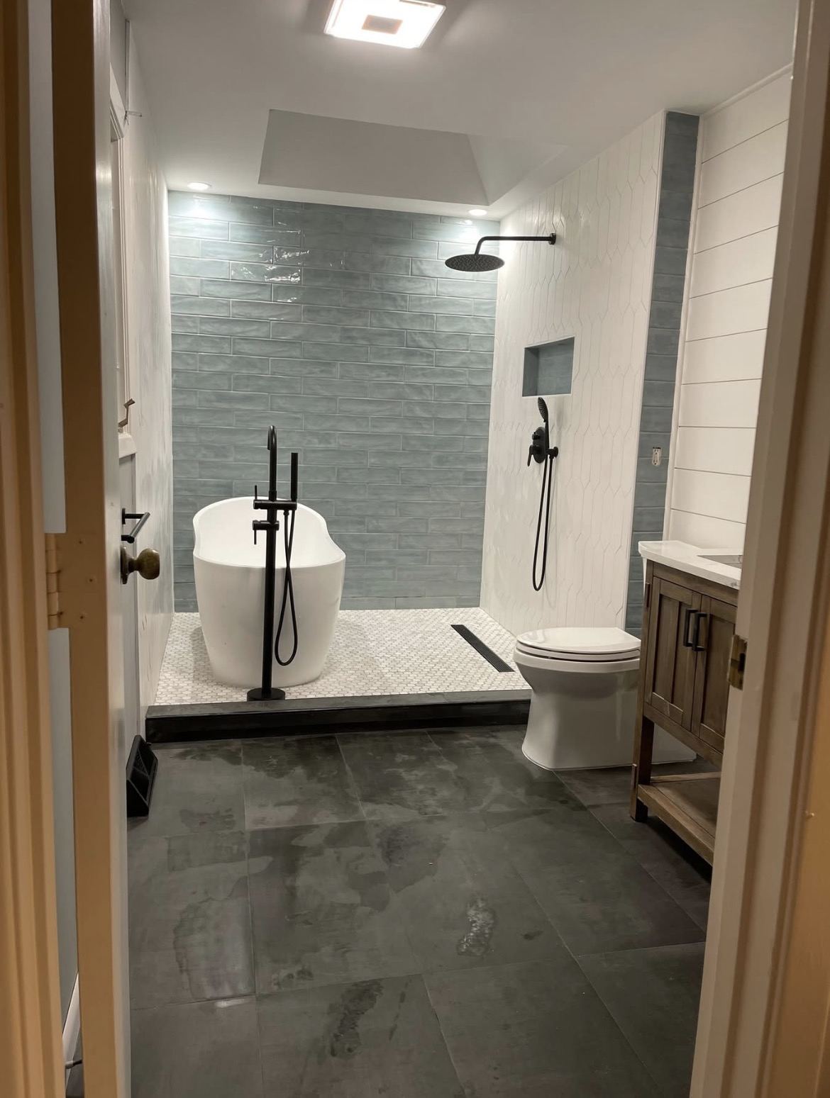 Renovated wet room with freestanding soaking tub, rainfall shower, linear drain, blue glass tile and matte black fixtures.