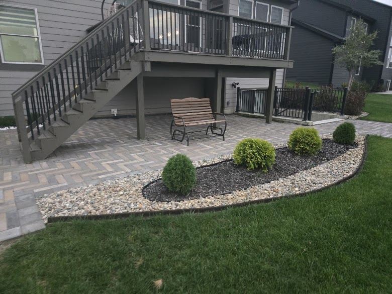 Paver patio beneath an elevated deck with a curved landscape bed, river rock, mulch and shrubs.